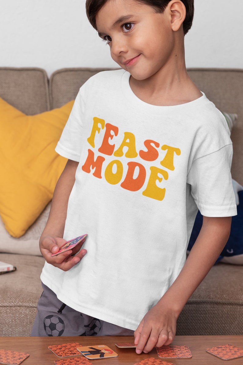 May include: White t-shirt with the words "FEAST MODE" in orange and yellow retro font. The shirt is worn by a child. The child is holding a card and is sitting at a wooden table with other cards.