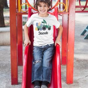 May include: White t-shirt featuring a green tractor design with Easter eggs and the name "Jacob" in black. The shirt is worn by a child on a red slide at a playground. The design includes blue, yellow, and green colors.