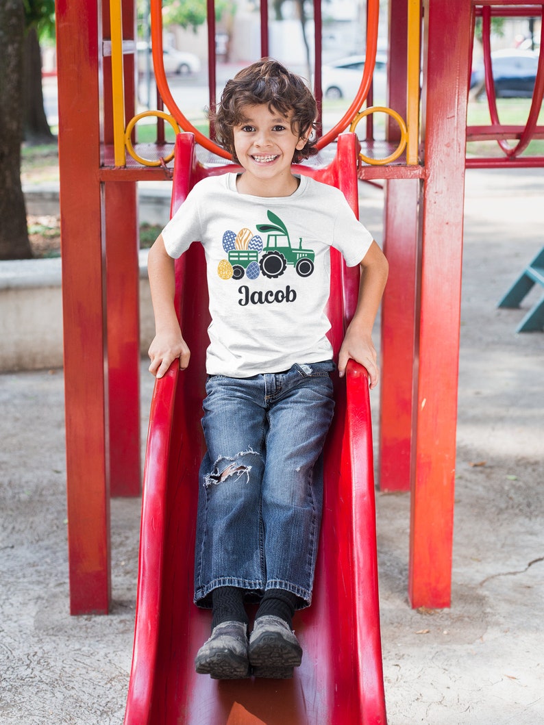 May include: White t-shirt featuring a green tractor design with Easter eggs and the name "Jacob" in black. The shirt is worn by a child on a red slide at a playground. The design includes blue, yellow, and green colors.