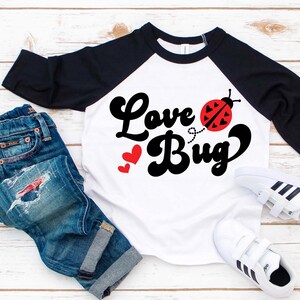 May include: White and black raglan style shirt with the words "Love Bug" in black script and a red ladybug graphic. The shirt is paired with blue jeans and white sneakers.
