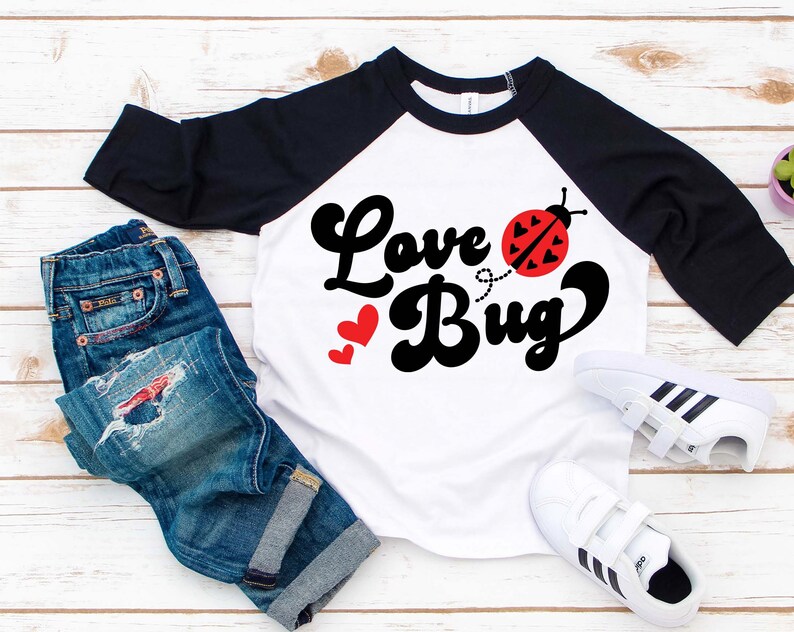 May include: White and black raglan style shirt with the words "Love Bug" in black script and a red ladybug graphic. The shirt is paired with blue jeans and white sneakers.