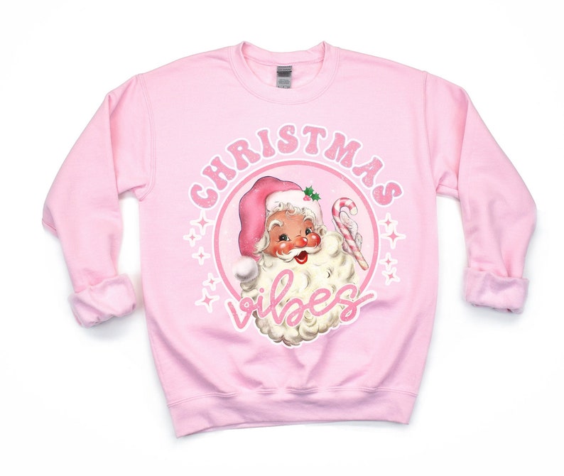 K&ouml;nnte beinhalten: Hellrosa Sweatshirt mit einem Vintage-Design von Santa Claus, der einen rosa Hut tr&auml;gt und eine Zuckerstange h&auml;lt. Die Worte "CHRISTMAS VIBES" sind in einem Kreis um Santa gedruckt.