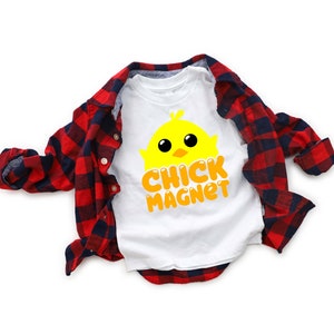 Puede incluir: Camiseta blanca con un gr&aacute;fico de pollito amarillo y las palabras "CHICK MAGNET" en naranja. Una camisa de franela a cuadros rojos y negros est&aacute; sobre la camiseta. El dise&ntilde;o es adecuado para Semana Santa o primavera.
