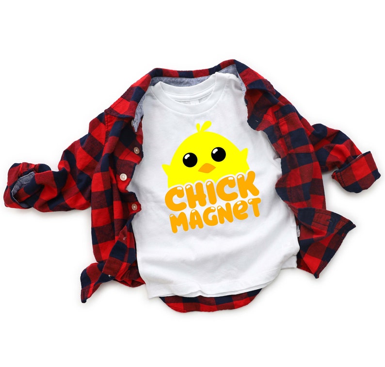 Puede incluir: Camiseta blanca con un gr&aacute;fico de pollito amarillo y las palabras "CHICK MAGNET" en naranja. Una camisa de franela a cuadros rojos y negros est&aacute; sobre la camiseta. El dise&ntilde;o es adecuado para Semana Santa o primavera.