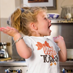 May include: A young child wearing a white t-shirt with a large orange bow and the words "little TURKEY" in brown and orange. The child has blonde hair and is smiling with their arms outstretched.