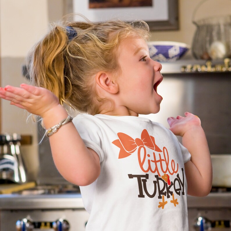 May include: A young child wearing a white t-shirt with a large orange bow and the words "little TURKEY" in brown and orange. The child has blonde hair and is smiling with their arms outstretched.