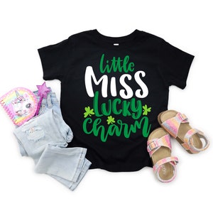 Puede incluir: Camiseta negra con el texto "little MISS lucky charm" en blanco y verde. La imagen incluye unos vaqueros azul claro, un cuaderno de unicornio rosa, una varita brillante y un par de sandalias brillantes.