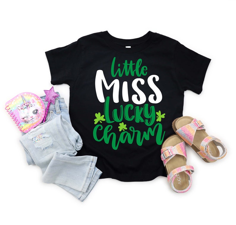 Puede incluir: Camiseta negra con el texto "little MISS lucky charm" en blanco y verde. La imagen incluye unos vaqueros azul claro, un cuaderno de unicornio rosa, una varita brillante y un par de sandalias brillantes.