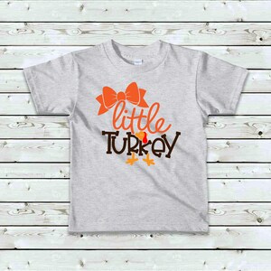 May include: A light gray t-shirt featuring the words "little TURKEY" in orange and brown, with an orange bow above the word "little". The design includes turkey feet. A festive shirt for Thanksgiving.