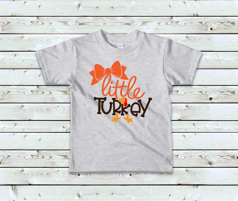 May include: A light gray t-shirt featuring the words "little TURKEY" in orange and brown, with an orange bow above the word "little". The design includes turkey feet. A festive shirt for Thanksgiving.