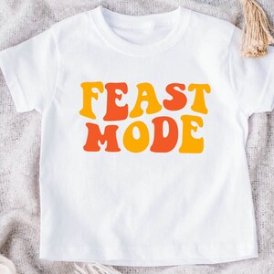 May include: White t-shirt with the words "FEAST MODE" in orange and yellow retro font. The shirt is a short-sleeved crew neck style, suitable for children. The design is centered on the front of the shirt.