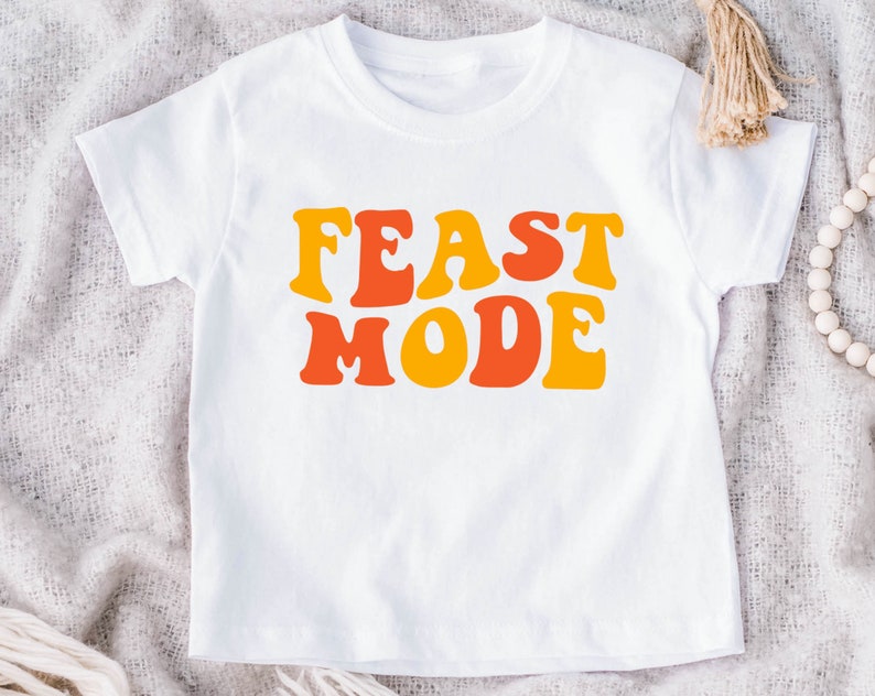 May include: White t-shirt with the words "FEAST MODE" in orange and yellow retro font. The shirt is a short-sleeved crew neck style, suitable for children. The design is centered on the front of the shirt.