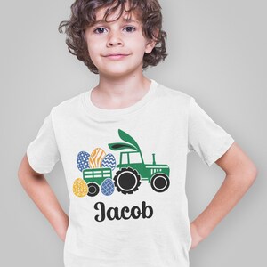 May include: White t-shirt featuring a green tractor pulling a wagon filled with Easter eggs. The eggs are decorated with various patterns and colors. The name "Jacob" is printed in black script below the tractor.