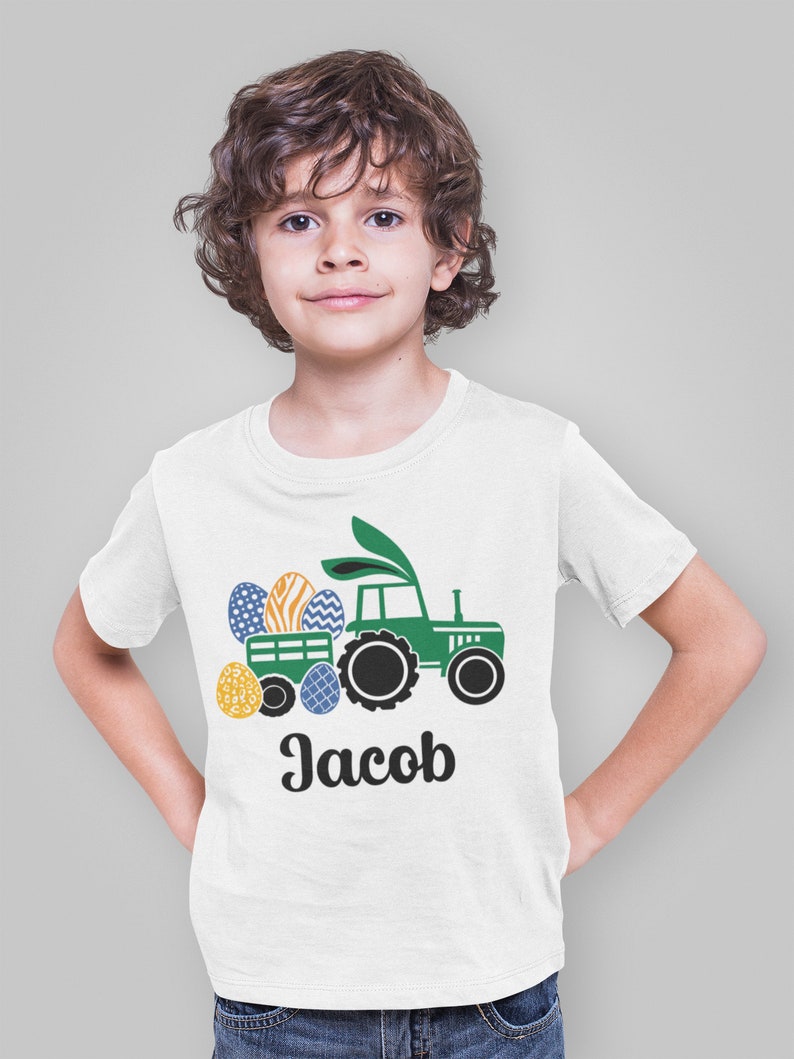 May include: White t-shirt featuring a green tractor pulling a wagon filled with Easter eggs. The eggs are decorated with various patterns and colors. The name "Jacob" is printed in black script below the tractor.