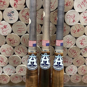 May include: Three wooden baseball bats with a dark finish, each featuring an American flag sticker and a logo. The bats have a signature and text that reads "PRO 11-7 | 33 MAPLE". The background shows a stack of wooden logs with handwritten numbers.