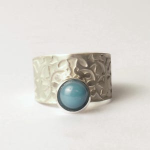 Könnte beinhalten: Ein breiter Silberring mit strukturierter Oberfläche und einem runden, hellblauen Stein. Der Ring hat ein gehämmertes Metallaussehen mit einer glatten, polierten Lünettenfassung für den Stein. Der Ring ist vor einem weißen Hintergrund platziert.