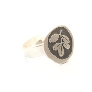 Puede incluir: Un anillo de plata con una parte superior ovalada texturizada que presenta un diseño detallado de una rama con hojas y bayas.