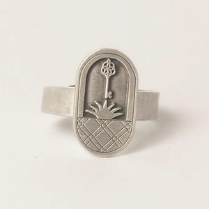 Può includere: Un anello d'argento con un design ovale che presenta una chiave con una testa a forma di fiore, una pianta e uno schema a scacchi.