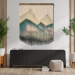 May include: A beige and white macrame wall hanging with a mountain range design. The mountains are made of yarn and have a blue and green ombre effect.