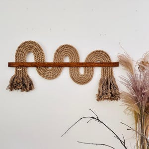 May include: A wall hanging made of natural rope woven into a wavy pattern with a wooden dowel across the top. The rope has a fringe on each end.