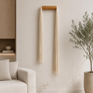 May include: A minimalist wall hanging with a natural wood bar and cream-colored fringe. The decor piece is mounted on a neutral-toned wall, complementing the modern living room setting. The room also features a beige sofa, a small vase, and a potted olive tree.