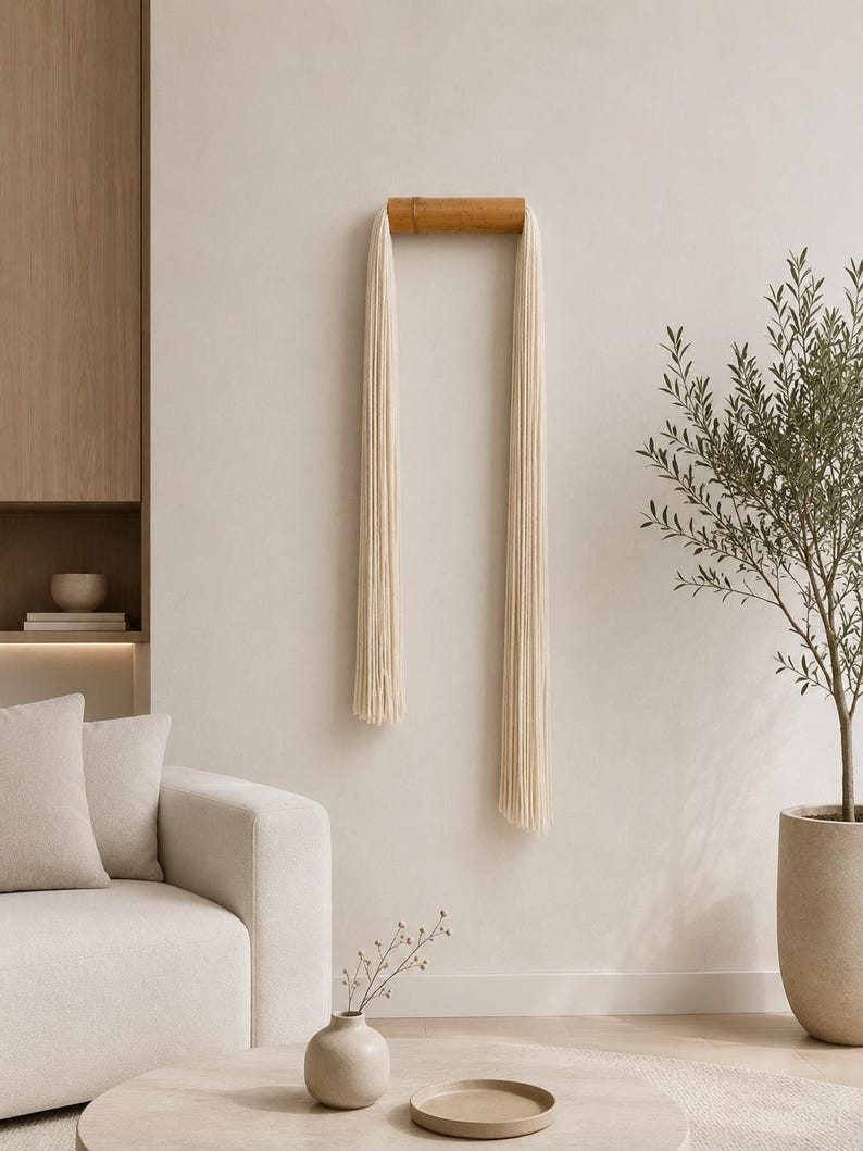 May include: A minimalist wall hanging with a natural wood bar and cream-colored fringe. The decor piece is mounted on a neutral-toned wall, complementing the modern living room setting. The room also features a beige sofa, a small vase, and a potted olive tree.