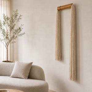 May include: A decorative wall hanging featuring a natural wood dowel with long, cream-colored fringe. The minimalist design is mounted on a neutral-toned wall, complementing the room's soft, modern aesthetic. The piece adds texture and a touch of bohemian style.
