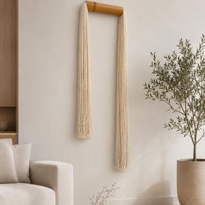 May include: A wall hanging featuring a natural bamboo frame with two long strands of cream-colored yarn. The minimalist design adds a touch of bohemian style to any room. The wall decor is a unique piece of art.
