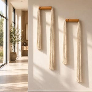 May include: Two wall-mounted macrame pieces. Each features a natural wood dowel with long, cream-colored fringe hanging below. The pieces are mounted on a neutral-toned wall, creating a minimalist, bohemian aesthetic. The room has a neutral color palette.