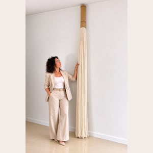 May include: A cream macrame wall hanging with a bamboo top is fixed to a white wall. The macrame features many vertical strands. A person in a beige pantsuit stands close by, gazing upwards at the artwork.