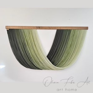 May include: A wall hanging made of green and black yarn, woven into a V-shape. The yarn is attached to a wooden dowel rod. The wall hanging is titled "Olivia Fiber Art art home".