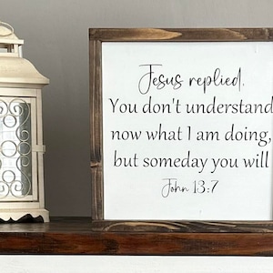May include: A wooden framed sign with a white background and black text. The text reads "Jesus replied. You don't understand now what I am doing, but someday you will. John 13:7".
