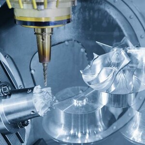 May include: A close-up of a CNC machine cutting a metal part. The machine has a drill bit attached to a robotic arm, and the part is being held in a vise. The machine is in a factory setting.