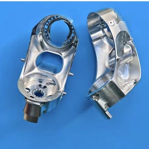 May include: Two polished, metallic silver-colored machine parts are displayed against a blue background. The parts have a complex, curved design with various openings and mounting points, suggesting they are components of a larger mechanical system.