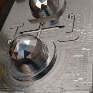 May include: Close-up of a metal mold with two round, faceted impressions. The mold is made of a silver-colored metal and has a textured surface.