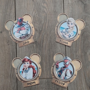 K&ouml;nnte beinhalten: Vier kreisf&ouml;rmige Wandplaketten in Form von Mickey-Maus-Ohren, die jeweils eine Figur in Matrosenkleidung zeigen: Deadpool mit der Aufschrift DAD, Elsa mit ELINA, Ariel mit MOM und Spider-Man mit TEDDY. Jede Plakette hat ein goldenes Finish.