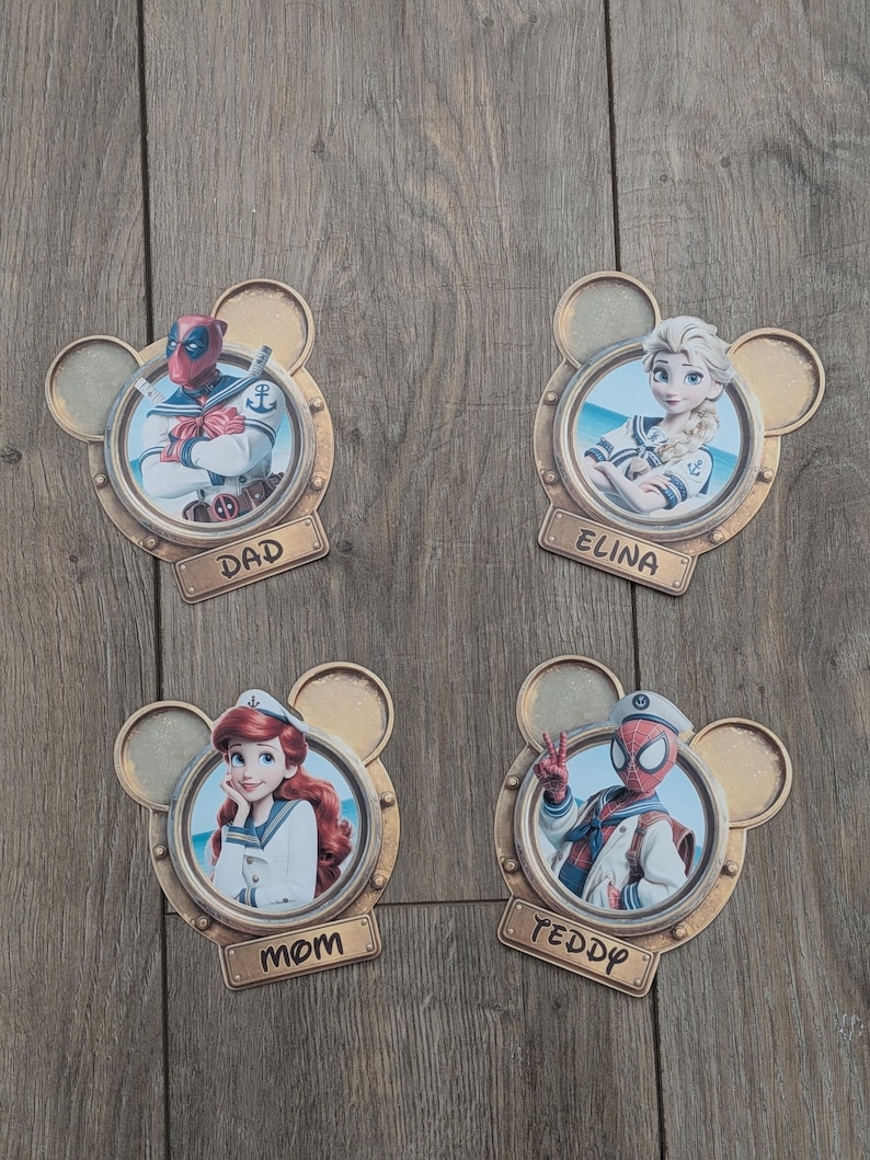 K&ouml;nnte beinhalten: Vier kreisf&ouml;rmige Wandplaketten in Form von Mickey-Maus-Ohren, die jeweils eine Figur in Matrosenkleidung zeigen: Deadpool mit der Aufschrift DAD, Elsa mit ELINA, Ariel mit MOM und Spider-Man mit TEDDY. Jede Plakette hat ein goldenes Finish.
