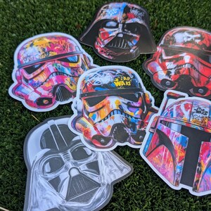 May include: Five colorful Star Wars character helmet stickers with graffiti designs. The stickers feature Darth Vader, a Stormtrooper, Boba Fett, and two different Stormtroopers. The stickers are all on a green grassy surface.