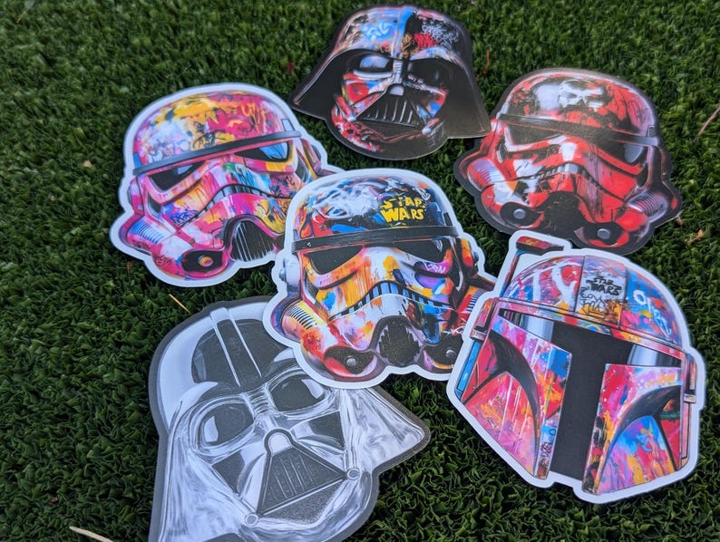 May include: Five colorful Star Wars character helmet stickers with graffiti designs. The stickers feature Darth Vader, a Stormtrooper, Boba Fett, and two different Stormtroopers. The stickers are all on a green grassy surface.