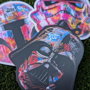 May include: Three colorful Star Wars character stickers with a graffiti design. The stickers feature Darth Vader, Boba Fett, and a Stormtrooper.