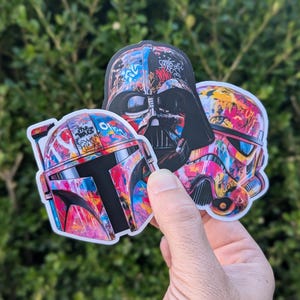 May include: Three Star Wars character stickers with a graffiti-style design. The stickers feature Darth Vader, Boba Fett, and a Stormtrooper. The stickers are colorful and have a variety of colors, including red, blue, green, yellow, and black.