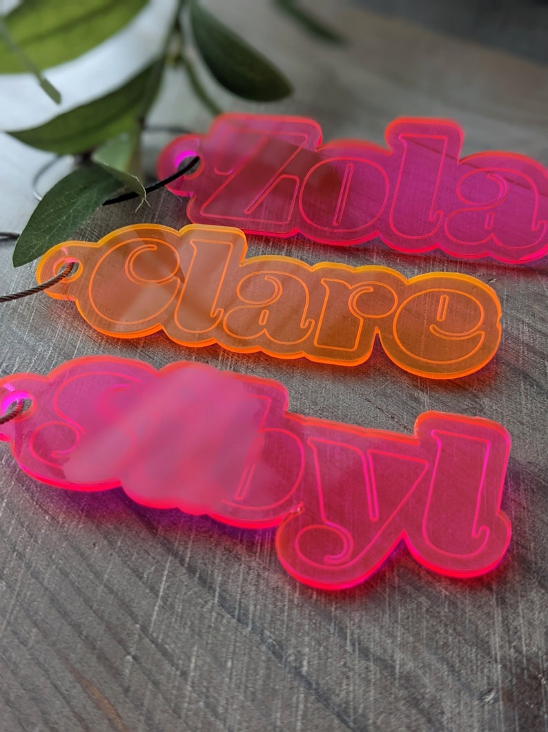May include: Three acrylic keychains with the names "Zola", "Clare", and "Saby" in a retro font. The keychains are pink and orange.