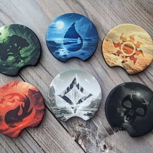May include: Six round coasters with different designs. Each coaster features a different element: a tree, a water drop, a sun, a mountain, a flame, and a skull. The coasters are made of a dark material and have a raised edge.