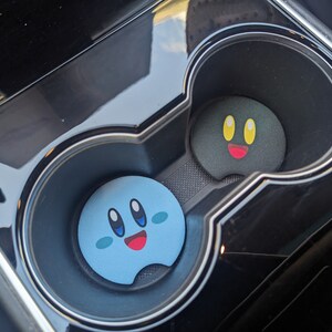 Peut inclure: Deux dessous de verre pour porte-gobelets de voiture. L'un est bleu clair avec un visage souriant, l'autre est noir avec un visage souriant. Les deux ont un design de dessin anim&eacute;. Les dessous de verre sont dans un porte-gobelet de console de voiture noire.