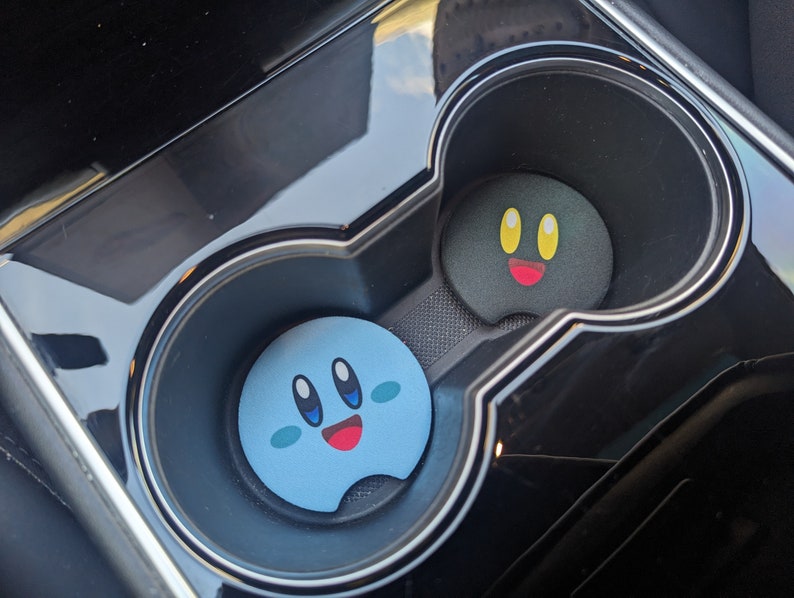 Peut inclure: Deux dessous de verre pour porte-gobelets de voiture. L'un est bleu clair avec un visage souriant, l'autre est noir avec un visage souriant. Les deux ont un design de dessin anim&eacute;. Les dessous de verre sont dans un porte-gobelet de console de voiture noire.
