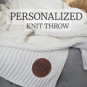 May include: A cream-colored knit throw blanket with the words "PERSONALIZED KNIT THROW" in a bold font. A brown leather patch with the initials "MIR" and the date "9/27/2024" is sewn on the blanket. The blanket is folded and draped.