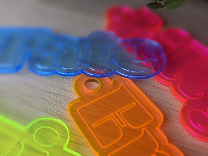 May include: Four colorful acrylic keychains with different designs. The keychains are blue, orange, green, and pink. The blue keychain says "Hello" in a bubbly font. The orange keychain has a simple design with a circle in the middle. The green keychain has a simple design with a circle in the middle. The pink keychain has a simple design with a circle in the middle.