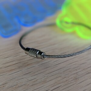 May include: A silver key chain with a braided cable and a silver clasp. The cable is looped around a wooden surface.
