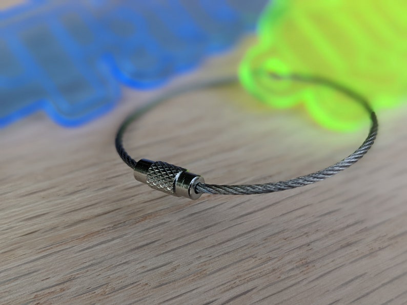 May include: A silver key chain with a braided cable and a silver clasp. The cable is looped around a wooden surface.