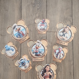 K&ouml;nnte beinhalten: Eine Sammlung von acht runden Wandplaketten in Form von Mickey-Maus-Ohren, die jeweils eine andere Figur in einem Bullaugenrahmen zeigen. Die Figuren umfassen Donald Duck, Deadpool, Alice, Sulley, Daisy Duck, Spider-Man, Stitch und Moana, mit Namensschildern f&uuml;r Jeff, Levi, Kylie, Josiah, Shauna, Isaiah, Lydia und Katie.
